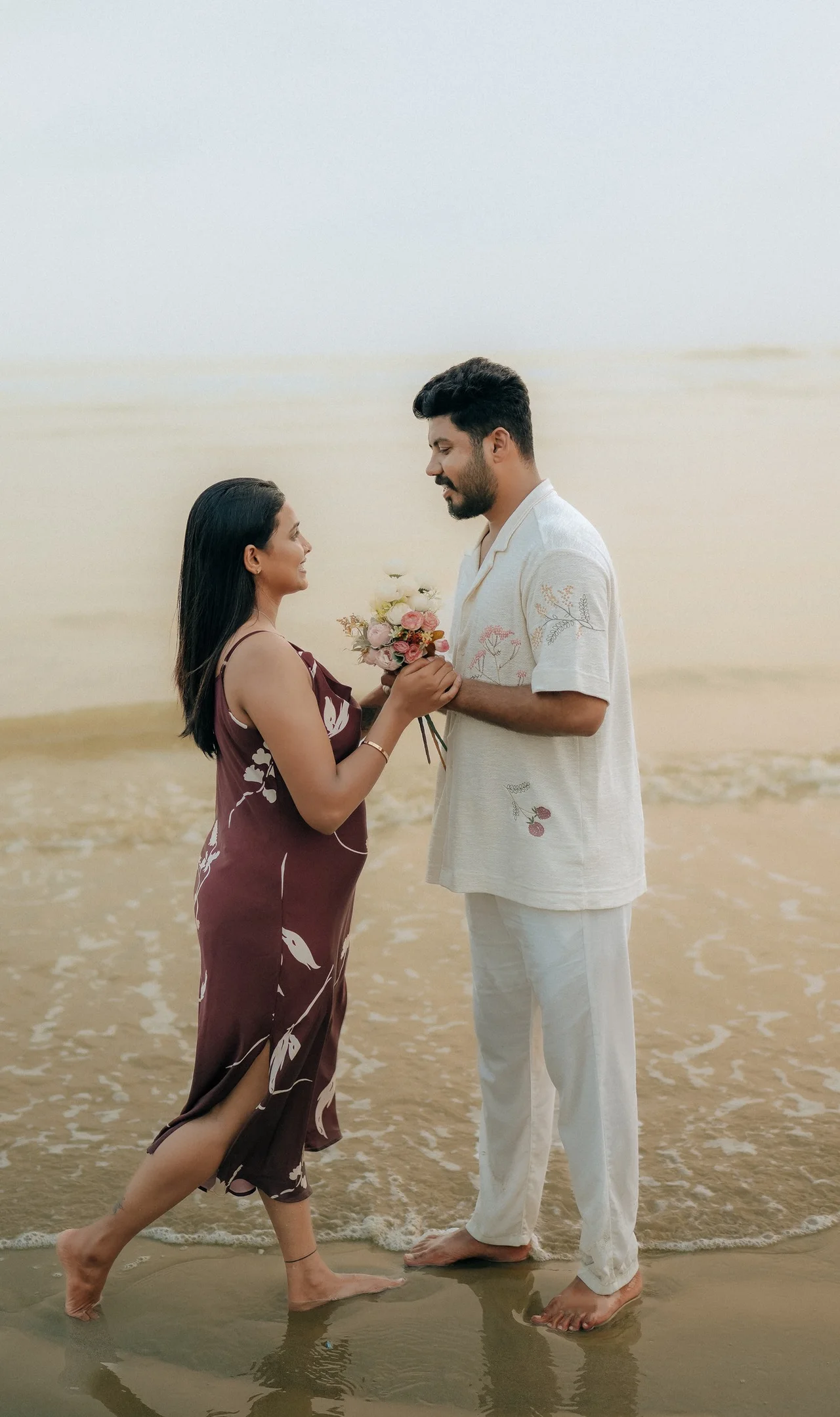 A couple facing each other at the water's edge on a misty beach, holding a small bouquet between them