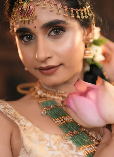 A close portrait of a bride in gold temple jewellery and a green bead necklace, holding a pink lotus
