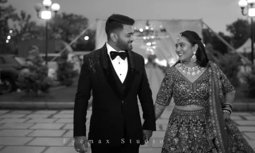 A black-and-white photo of a couple walking hand in hand past lit lamps, he in a tuxedo and bow tie, she in an embroidered lehenga, smiling at each other