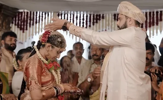 A groom in a cream turban rests his hand on the bowed head of his bride, in an orange silk saree with roses and jasmine in her hair, as guests look on at their wedding