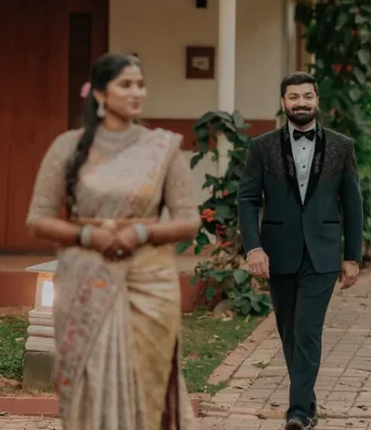 A groom in a black tuxedo walks forward smiling, while a woman in a saree stands in soft focus in front of him