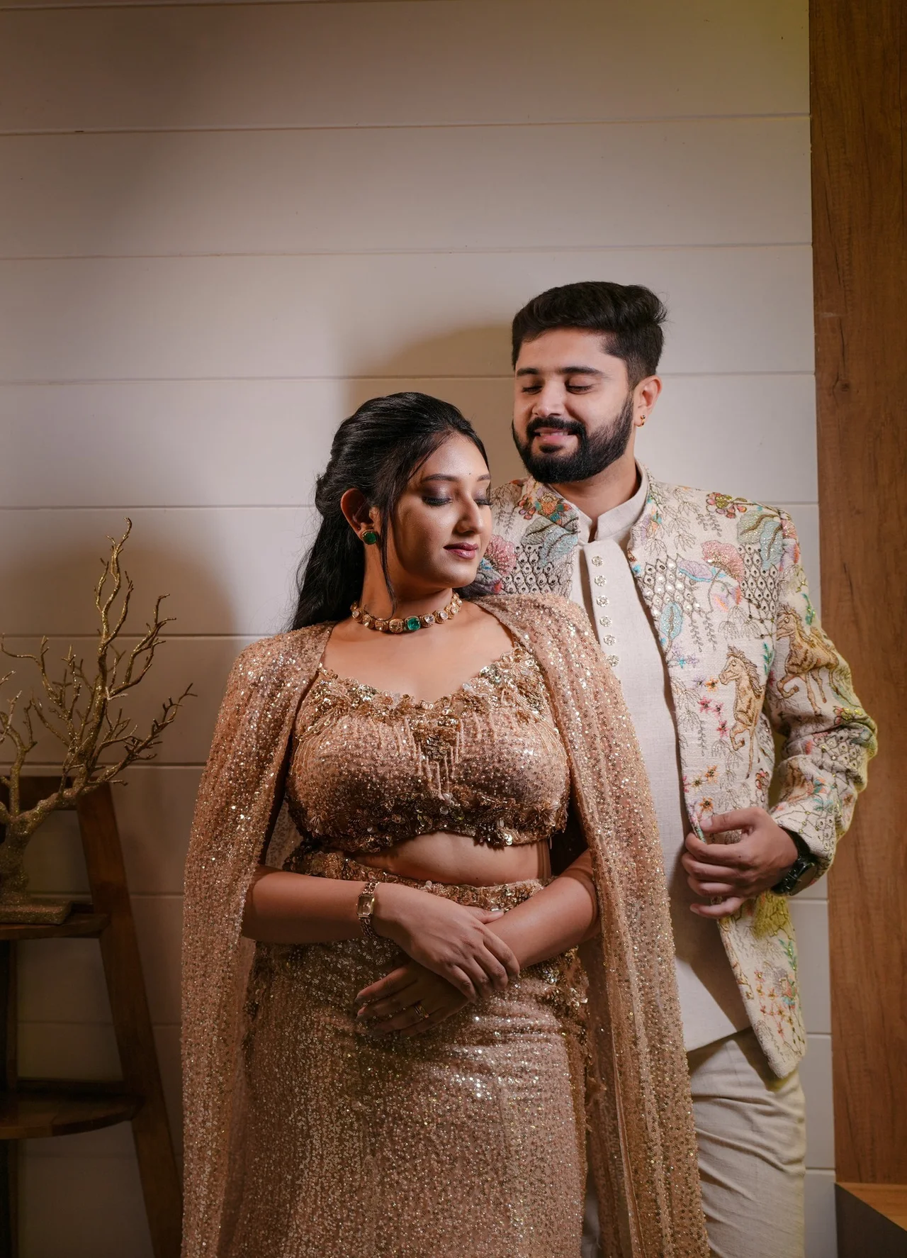 A couple standing close against a white panelled wall, she in a sequinned blush lehenga and cape, he in an embroidered jacket