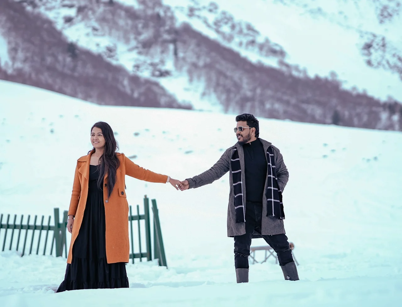 A couple holding hands in deep snow below a mountain, she in an orange coat, he in a grey coat and checked scarf