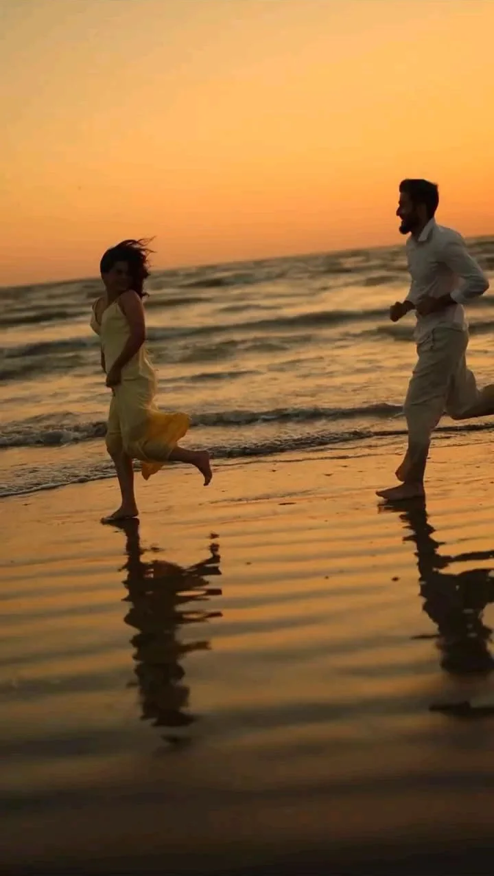 A couple running barefoot along the shore at sunset, their reflections on the wet sand
