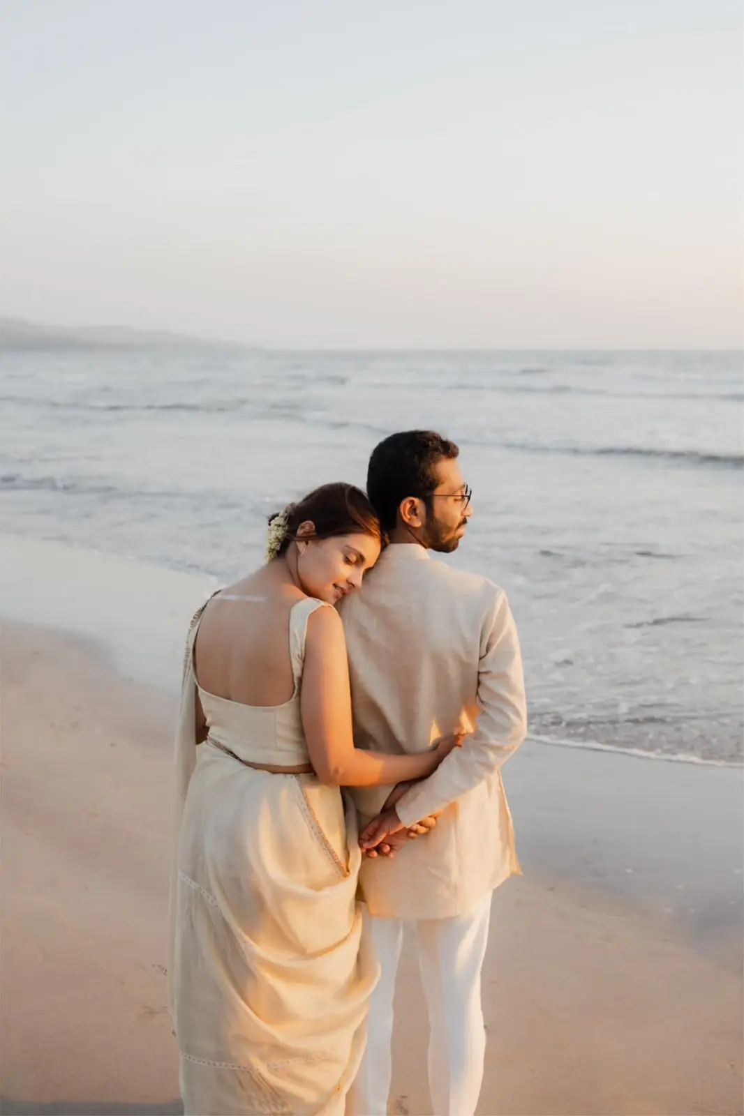A couple on a beach in soft golden light, seen from behind, her head resting on his shoulder